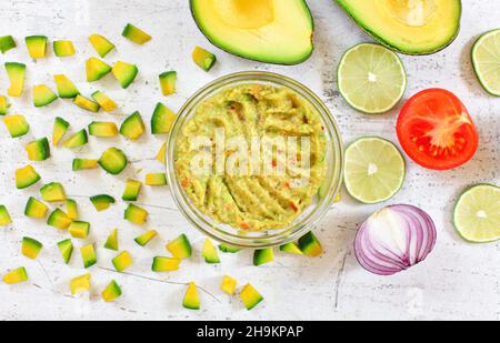 Avocado Hälften und Stücke, Limetten Tomaten, Zwiebeln - grundlegende Guacamole Zutaten auf weißem Brett, fertig mischen in einer kleinen Glasschüssel in der Nähe Stockfoto