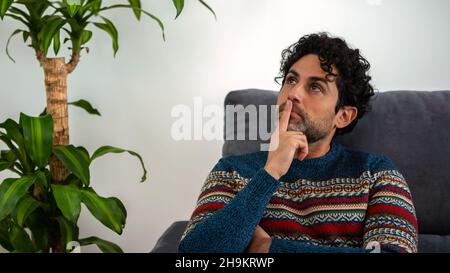 Gesicht eines nachdenklichen erwachsenen Mannes, der nachdenklich aufschaute, während er auf der Couch zu Hause saß. Bärtiger Mann, der die Wahl getroffen hat oder auf einem Sofa nachdenkt und wegschaut. Stockfoto