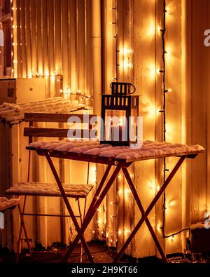 Ein Holztisch und ein mit Schnee bedeckter Stuhl, beleuchtet mit Weihnachtslichtern. Eine dekorative Kerzenlaterne brennt auf dem Tisch. Winternacht. Stockfoto