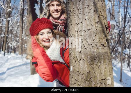 Verspieltes Paar versteckt sich hinter einen Baum im Schnee Stockfoto