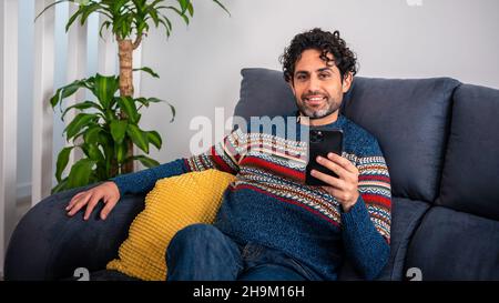 Glücklicher lächelnder Mann mit Smartphone-Gerät, während er zu Hause auf dem Sofa sitzt. Reifer Mann, der auf der Couch liegt und Nachrichten auf dem Mobiltelefon liest. kaukasisches spanisch Stockfoto