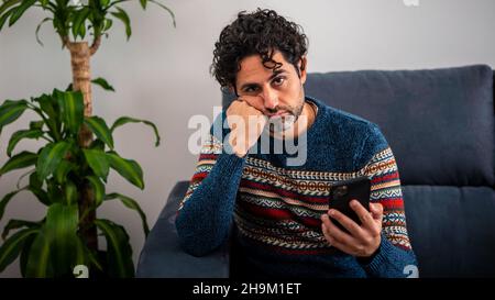 Männliche Depression, die seine Stirn hält, während sie Kopfschmerzen auf der Couch zu Hause hat. Erwachsener Mann mit Smartphone und Denken, mit schweren Gedanken, breake up w Stockfoto