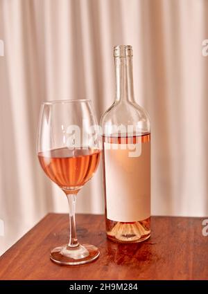 Flasche Roséwein mit leerem Etikett mit Glas auf Holztisch mit weißem Vorhang im Hintergrund. Vertikal. Weinprobe in Pink Stockfoto