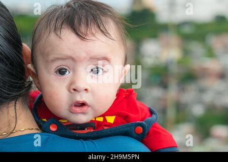 Schönes lateinisches Baby, das seinen Kopf über die Schulter seiner Mutter blickt, großes Baby mit erstauntem Gesicht, blauem Overall und rotem Hemd, das zurückblickt. Mit dem Mund Stockfoto