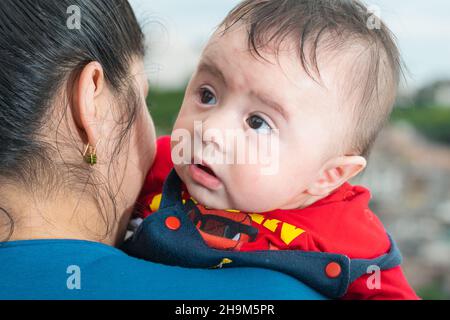 Schönes lateinisches Baby, das seinen Kopf über die Schulter seiner Mutter blickt, großes Baby mit den Augen zum Himmel, blaue Overalls und rotes Hemd. Mit offenem Mund Stockfoto