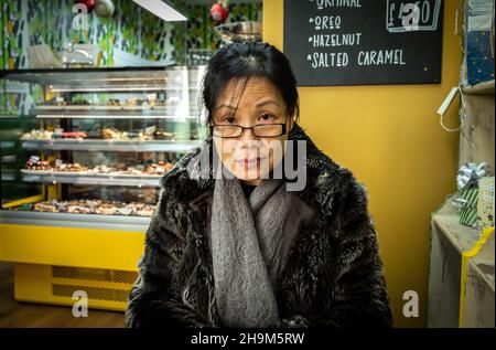 Eine Vietnamesin mit einem Kunstpelzmantel und Schal sitzt in einem handgefertigten Schokoladencafé namens Cenu Cacao in King's Parade, Cambridge, Großbritannien Stockfoto