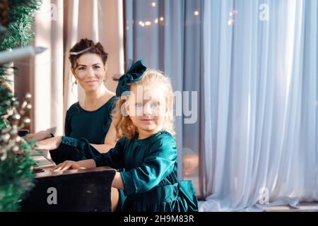 Portrait eines kleinen Mädchens in niedlichem grünen Kleid, das Klavier spielt, verziert mit Weihnachtsornamenten. Glückliche Mutter lächelt auf dem Hintergrund. Winter- und Neujahrsferien. Kopieren Sie die Spase. Stockfoto