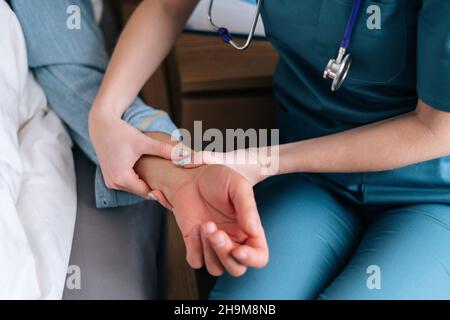 Nahaufnahme einer nicht erkennbaren Krankenschwester, die den Puls am Handgelenk überprüft und einen kranken älteren männlichen Patienten auf dem Bett im Krankenhauszimmer anliegt. Stockfoto