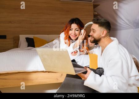 Foto eines jungen Paares in Bademänteln, das mit einem Laptop auf dem Bett liegt. Mann und Frau hören Musik. Stockfoto