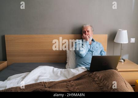 Nachdenklicher grauhaariger älterer Mann mit Bart, der einen Laptop-Computer benutzt, der zu Hause auf einem bequemen Bett liegt. Stockfoto