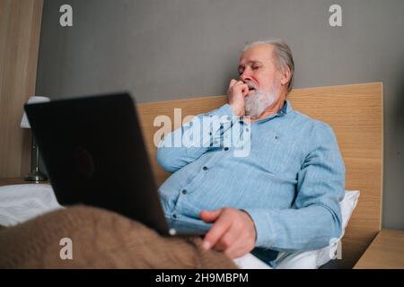 Low-Angle-Ansicht eines frustrierten grauhaarigen älteren Mannes mit Bart mit Laptop-Computer, der zu Hause auf dem Bett sitzt. Stockfoto