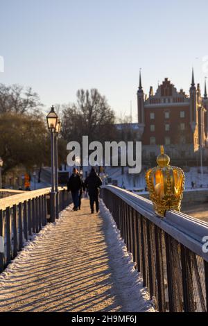 Stockholm im Winter. Menschen, die auf der Brücke mit einer vergoldeten Krone auf Skeppsholmsbron mit Strandvagen im Hintergrund laufen. Stockfoto