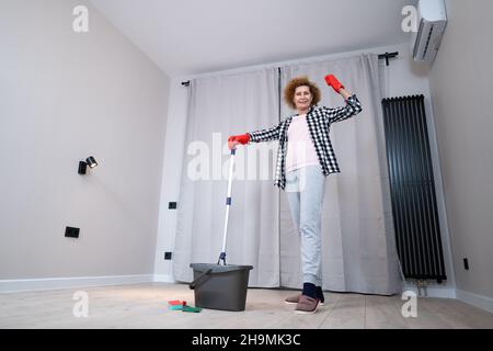 Menschen-, Hausarbeit- und Haushaltskonzept. Glückliche ältere Frau in Schutzhandschuhen reinigt den Boden und tanzt zu Hause in einer leeren Wohnung, bevor sie sich bewegt Stockfoto
