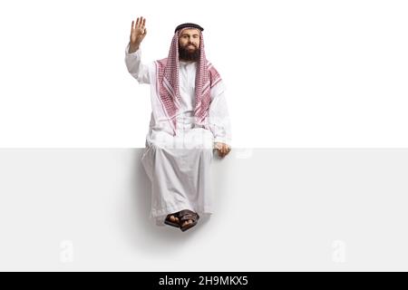 Saudi-arabischer Mann, der auf einer leeren Tafel sitzt und isoliert auf weißem Hintergrund auf die Kamera zuwinkt Stockfoto