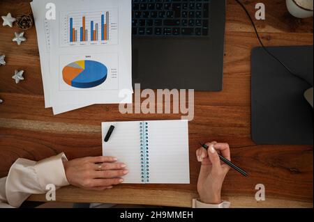 Draufsicht einer Geschäftsfrau, die arbeitete, aß ihren Schreibtisch und schrieb auf einem Notizblock, während sie statistische Berichte und Diagramme durchläuft. Stockfoto