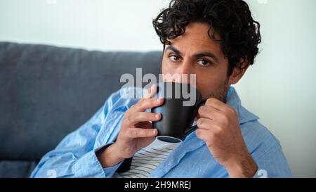 Kaukasischer Mann mit Bart, der morgens Espresso trinkt und auf die Kamera schaut, während er auf der Couch sitzt. Schönes männliches Modell. Stockfoto