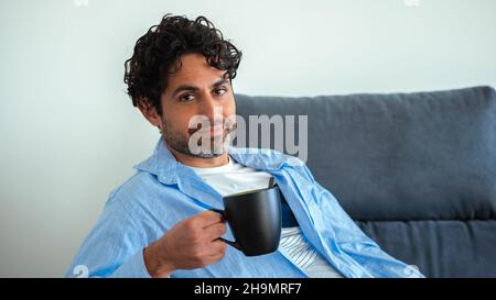 Hübscher Mann mit Bart trinkt morgens Espresso-Kaffee mit glücklichem Gesicht lächelnd und schaut auf die Kamera auf dem Sofa von zu Hause. Positive Person. Stockfoto