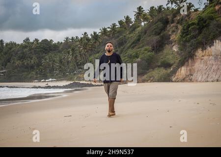 Ein junger Mann mit Hippie-Aussehen, der an einem einsamen Strand spazierengeht. Einsamkeit und Depression Konzept. Stockfoto