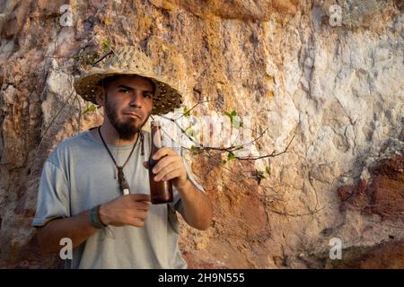 Junger Hippie-Reisender in einem Strohhut mit einer Flasche Bier neben einigen riesigen Felsen, der auf die Kamera schaute. Stockfoto