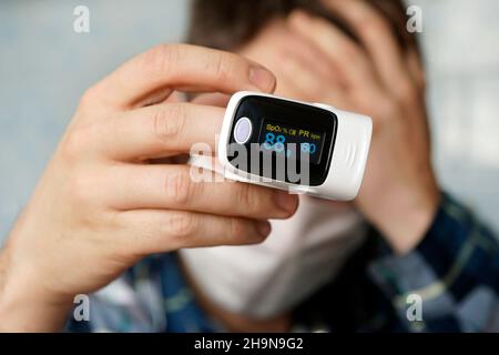 Ein Patient in einer medizinischen Maske misst die Blutsättigung mit einem elektronischen Pulsoximeter an seinem Finger. Pulsoximeter zur Messung der Pulsfrequenz und Stockfoto