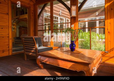 Sofa-Stuhl aus Holz mit grauen Kissen, indonesischer Teakholz-Couchtisch im Inneren des Sonnenzimmers mit Holzrahmen und Blick auf den Teich und den Hinterhof. Stockfoto