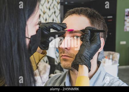 Mann in einem Schönheitssalon während der Augenbrauenanpassung. Eine Frau entfernt überschüssige Haare auf dem Gesicht eines Mannes. Stockfoto