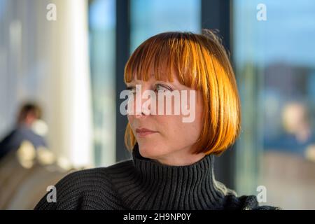 Porträt einer attraktiven trendigen Frau mittleren Alters mit rothaarigen Haaren, die drinnen sitzt und links vom Rahmen in einer Nahaufnahme von der Seite schaut Stockfoto