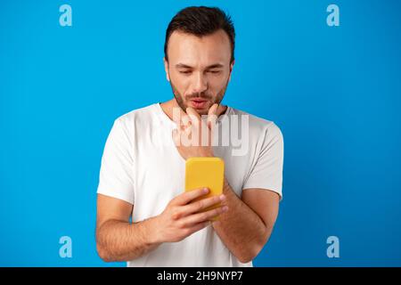 Staunender Kerl, der auf den Smartphone-Bildschirm in Schock über blauem Hintergrund schaut Stockfoto