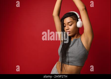 Selbstbewusst ruhige schöne afroamerikanische junge Frau mit kabellosen Kopfhörern in grauer Sportkleidung genießt Musik hören, tanzen mit erhobenen Armen a Stockfoto