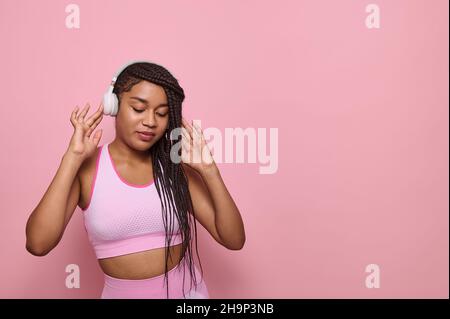 Hübsche, sportliche, schwarze Frau mit stylischen Afro-Pigtails, die Kopfhörer trägt, tanzt mit geschlossenen Augen auf einem rosa Hintergrund mit Platz für Kopien. Bsb Stockfoto