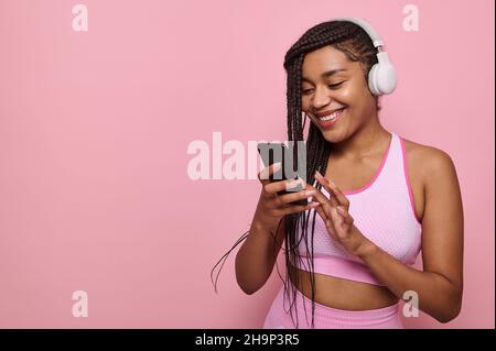 Attraktive afroamerikanische Sportlerin in rosa Sportbekleidung und kabellosen Kopfhörern durchsucht Webseiten im Internet im Smartphone, lächelt mit fröhlichem t Stockfoto