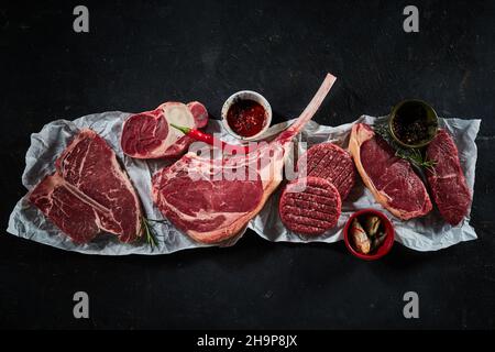 Draufsicht auf verschiedene rohe Rindersteaks, darunter Tomahawk-Sirloin mit verschiedenen Gewürzen, Chili und Rosmarinzweigen Stockfoto