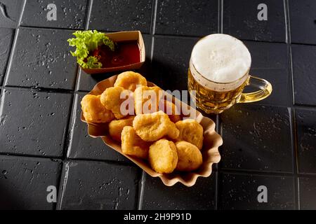 Hoher Winkel von leckeren heißen Chicken Nuggets und ein Glas alkoholisches Bier mit Schaum auf schwarzem Tisch platziert Stockfoto