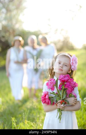 Kleines Mädchen in weißem Kleid halten Blumen, verschwommen Familie im Hintergrund. Stockfoto