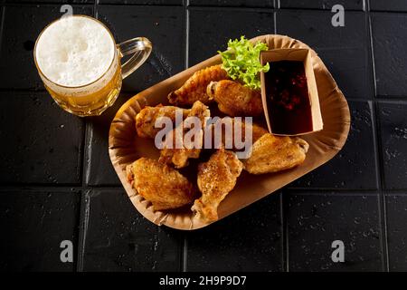 Von oben Teller mit gebratenen Hähnchenflügeln mit Sauce und Glas Bier auf schwarzen Tisch im Restaurant gelegt Stockfoto