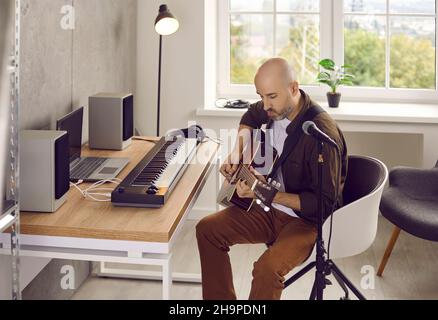 Fokussierter ernsthafter männlicher Musiker, der Akustikgitarre spielt und in seinem Heimstudio einen neuen Song kreiert. Stockfoto