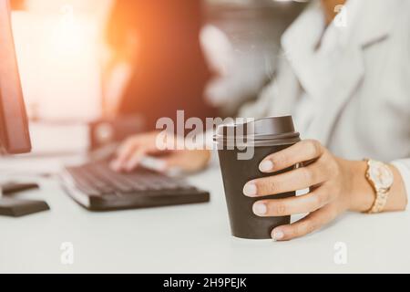 Hand an Kaffeetasse im Büroschreibtisch. Geschäftsleute trinken Kaffee aus Papiertasse für gute Arbeit Konzept Stockfoto