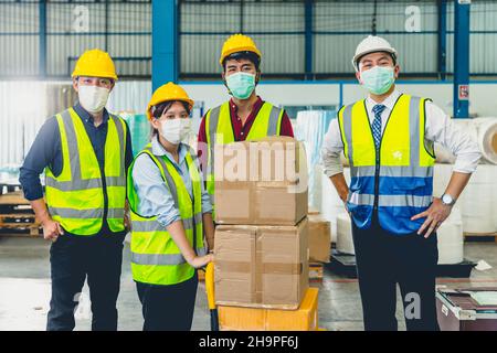 Eine Gruppe asiatischer Teamarbeiter trägt eine Gesichtsmaske, die im Lagerhaus zusammensteht Stockfoto