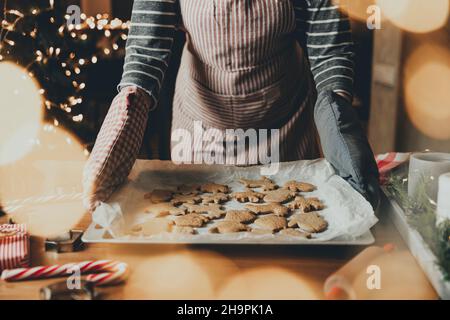 Frohe Weihnachten, Frohes neues Jahr. Lebkuchenkochen, Kuchen oder Strudelbacken. Eine junge Frau oder Mutter hält Deco und hält ein Blatt mit fertig gebackenen Keksen in ihren Handschuhen. Hochwertige Fotos Stockfoto