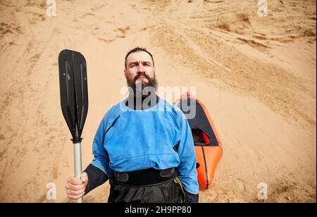 Porträt des alten Mannes mit schwarzem Bart und Paddel in blauer Jacke in der Nähe von orangefarbenen Floß Sportboot auf Sanddüne Hintergrund Stockfoto
