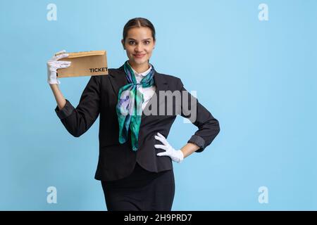 Halblanges Porträt eines schönen jungen Mädchens, Flugbegleiter in schwarzer Uniform posiert isoliert auf blauem Studiohintergrund. Stockfoto