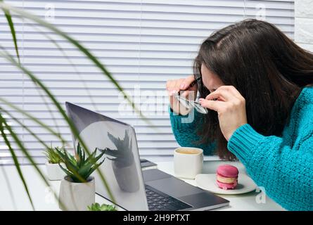 Eine Frau in einem blauen Pullover hält eine Brille in den Händen und blickt aufmerksam auf den Bildschirm. Videoanruf, überraschen Sie von dem, was Sie auf dem Bildschirm sehen Stockfoto