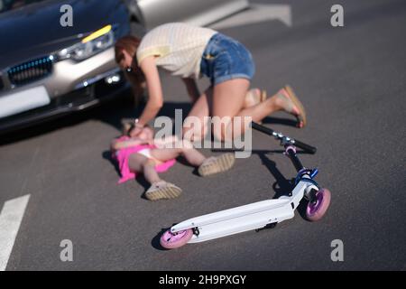 Kleines Mädchen liegt auf dem Asphalt mit Roller und ihre Mutter nach Kollision mit dem Auto Stockfoto