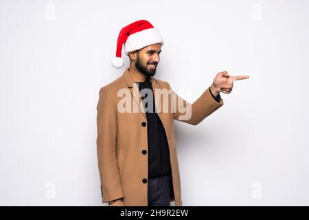 Der junge Mann im Weihnachtsmannhut ist überrascht und zeigt die Finger auf isolierten weißen Hintergrund Stockfoto