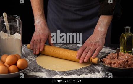 Ein Mann rollt den Teig mit einem Nudelholz für ein traditionelles russisches Gericht Knödel, Zutaten für den Teig und Hackfleisch auf dem Tisch aus Stockfoto