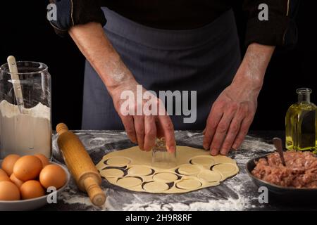 Ein Mann schneidet eine Form für ein traditionelles russisches Gericht Knödel, Zutaten für den Teig und Hackfleisch auf dem Tisch Stockfoto