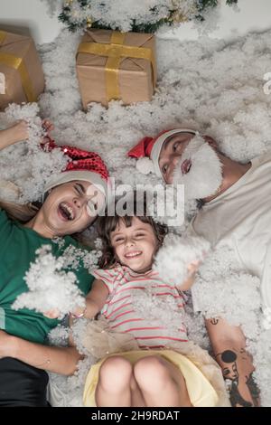 Weihnachtsfoto der glücklichen Familie. Neujahrsporträt von Vater, Mutter und Tochter in weihnachtlichen roten Hüten, lächelnd und im weißen Kunstschnee liegend. Stockfoto