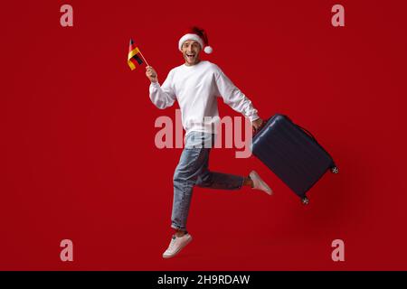 Fröhlicher Junger Mann Im Weihnachtsmütze-Springen Mit Deutscher Flagge Und Koffer Stockfoto