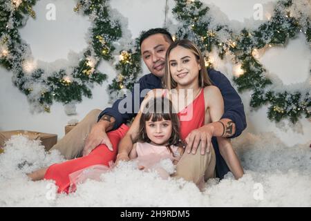 Weihnachts-Studio-Familiendreh. Neujahrsfoto der glücklichen Familie. Weihnachtskränze und künstlicher Schnee. Mutter, Vater und süße Tochter. Stockfoto