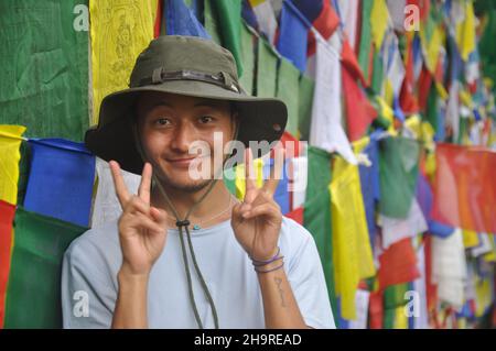 Foto eines glücklichen asiatischen jungen Mannes, der die Kamera anschaut, mit seinen Händen Friedenszeichen macht und sich gegen tibetisch-buddhistische Gebetsfahnen posiert Stockfoto